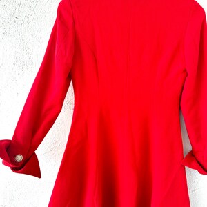 Puede incluir: Un blazer rojo brillante con mangas largas y una falda acampanada. Las mangas tienen pu&ntilde;os doblados con botones dorados. El blazer est&aacute; colgado en una percha blanca contra una pared blanca texturizada.