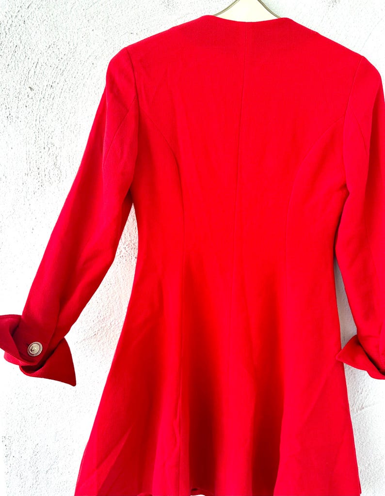 Puede incluir: Un blazer rojo brillante con mangas largas y una falda acampanada. Las mangas tienen pu&ntilde;os doblados con botones dorados. El blazer est&aacute; colgado en una percha blanca contra una pared blanca texturizada.