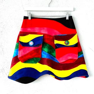 May include: A colorful mini skirt with a wavy pattern in shades of red, yellow, blue, green, and black. The skirt has two large pockets with button closures.