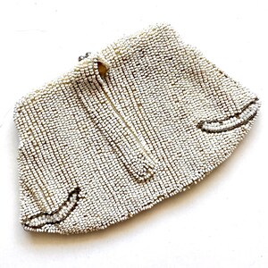May include: A white beaded clutch purse with a silver clasp. The purse is made of small, round beads that are sewn together in a close pattern. The purse has a curved shape and a pointed bottom.