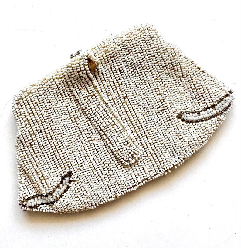May include: A white beaded clutch purse with a silver clasp. The purse is made of small, round beads that are sewn together in a close pattern. The purse has a curved shape and a pointed bottom.