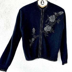 Puede incluir: Un c&aacute;rdigan negro con un dise&ntilde;o floral de cuentas. El c&aacute;rdigan tiene manga larga y cierre de botones.