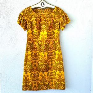 May include: A vintage yellow dress with a floral pattern in shades of brown and gold. The dress features short, ruffled sleeves and a square neckline. It is hanging on a vintage hanger.