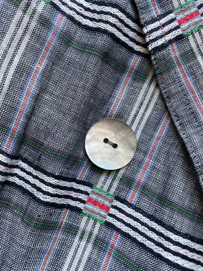 May include: A textured fabric garment featuring a dark gray base with a woven plaid pattern in white, red, green, and blue stripes. A single round, iridescent, two-hole button is centered on the fabric, secured with dark gray thread.