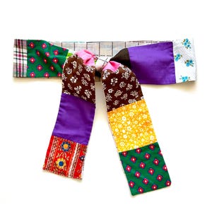 May include: A colorful patchwork fabric belt with a bow tie. The belt is made of different patterned fabrics in shades of brown, purple, green, yellow, red, and white.