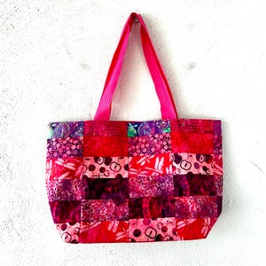 May include: A pink patchwork tote bag featuring a variety of floral and geometric patterns. The bag has two pink handles and is made of fabric.