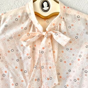 May include: A cream-colored blouse with a floral pattern in peach, black, and gray. The blouse features a bow tie at the neckline and is displayed on a vintage hanger with a cameo detail. The fabric appears lightweight and semi-sheer.