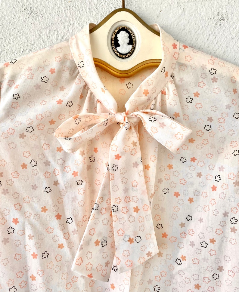 May include: A cream-colored blouse with a floral pattern in peach, black, and gray. The blouse features a bow tie at the neckline and is displayed on a vintage hanger with a cameo detail. The fabric appears lightweight and semi-sheer.