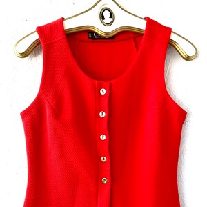 May include: A sleeveless, bright red dress with a textured fabric and a row of white buttons down the front. The dress is hanging on a white and gold hanger.