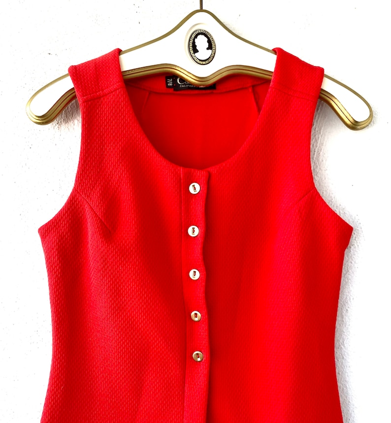 May include: A sleeveless, bright red dress with a textured fabric and a row of white buttons down the front. The dress is hanging on a white and gold hanger.