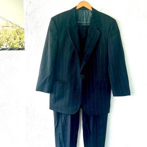 Peut inclure: Un costume bleu marine fonc&eacute; orn&eacute; d'un motif &agrave; fines rayures verticales. L'ensemble se compose d'une veste &agrave; simple boutonnage avec des revers crant&eacute;s, une fermeture &agrave; un bouton et deux poches avant, assortie &agrave; un pantalon habill&eacute;.