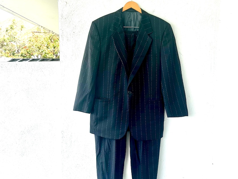 Peut inclure: Un costume bleu marine fonc&eacute; orn&eacute; d'un motif &agrave; fines rayures verticales. L'ensemble se compose d'une veste &agrave; simple boutonnage avec des revers crant&eacute;s, une fermeture &agrave; un bouton et deux poches avant, assortie &agrave; un pantalon habill&eacute;.