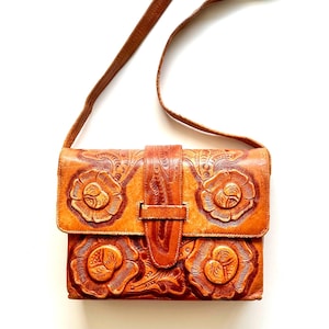 May include: A tan leather crossbody bag with a floral embossed design. The bag features a flap closure with a buckle and a long, thin strap. The floral pattern is detailed and raised, adding texture to the bag's surface.