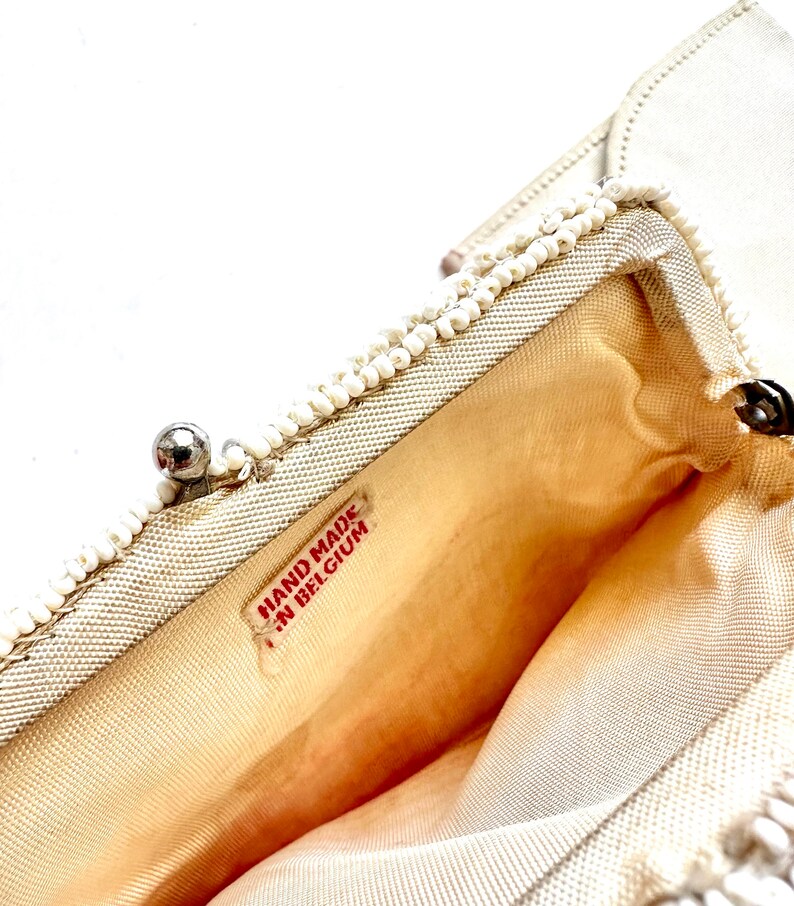 May include: White beaded clutch purse with a silver clasp and a red label that reads "HAND MADE IN BELGIUM".