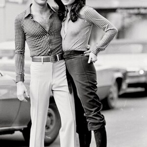 May include: A black and white photo of a couple standing in front of a car. The man is wearing a checkered shirt and white pants. The woman is wearing a checkered shirt, black pants, and black boots.