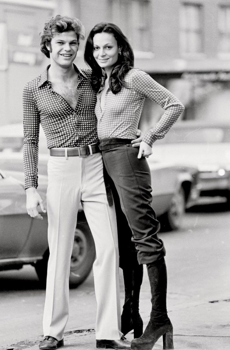 May include: A black and white photo of a couple standing in front of a car. The man is wearing a checkered shirt and white pants. The woman is wearing a checkered shirt, black pants, and black boots.