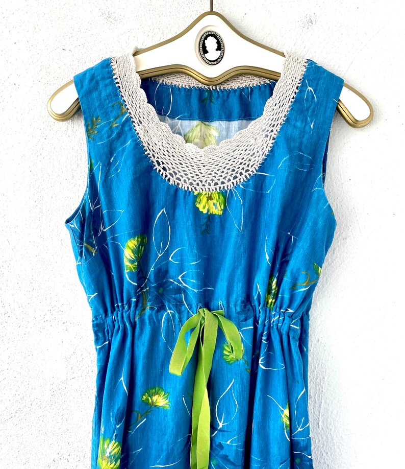 May include: A blue sleeveless dress with a white crochet neckline and a green ribbon tie at the waist. The dress has a floral print with white outlines.