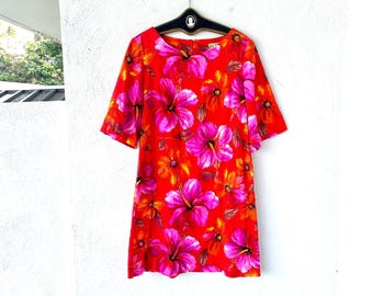 Könnte beinhalten: Ein leuchtend rotes Kleid mit einem Blumenmuster aus großen rosa und orangefarbenen Hibiskusblüten. Das Kleid hat kurze Ärmel und einen runden Ausschnitt. Es hängt an einem schwarzen Kleiderbügel vor einer weißen Wand.