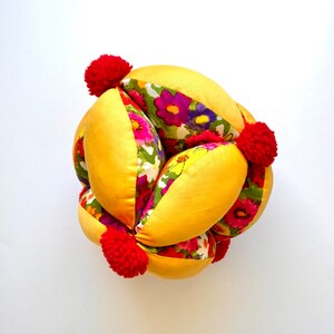 May include: A yellow fabric ball with a floral pattern and red pom-poms. The ball is made of multiple fabric pieces sewn together.