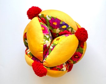 May include: A yellow fabric ball with a floral pattern and red pom-poms. The ball is made of multiple fabric pieces sewn together.