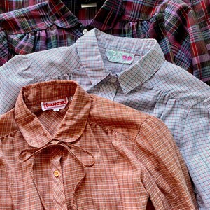 May include: Three plaid shirts stacked on top of each other. The top shirt is a purple and blue plaid, the middle shirt is a light blue and red plaid, and the bottom shirt is a brown and white plaid. The shirts have different labels on the collars, including "WELD X HUNTER", "HUK A P", and "FOXMOOR".