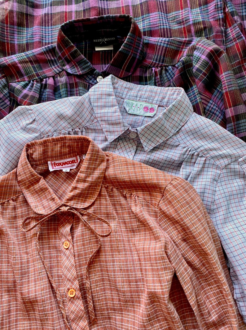 May include: Three plaid shirts stacked on top of each other. The top shirt is a purple and blue plaid, the middle shirt is a light blue and red plaid, and the bottom shirt is a brown and white plaid. The shirts have different labels on the collars, including "WELD X HUNTER", "HUK A P", and "FOXMOOR".