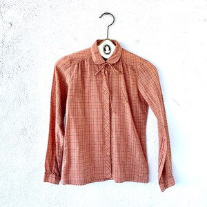 May include: A long-sleeved, button-down shirt with a plaid pattern in shades of pink and orange. The shirt has a Peter Pan collar and a bow tie.
