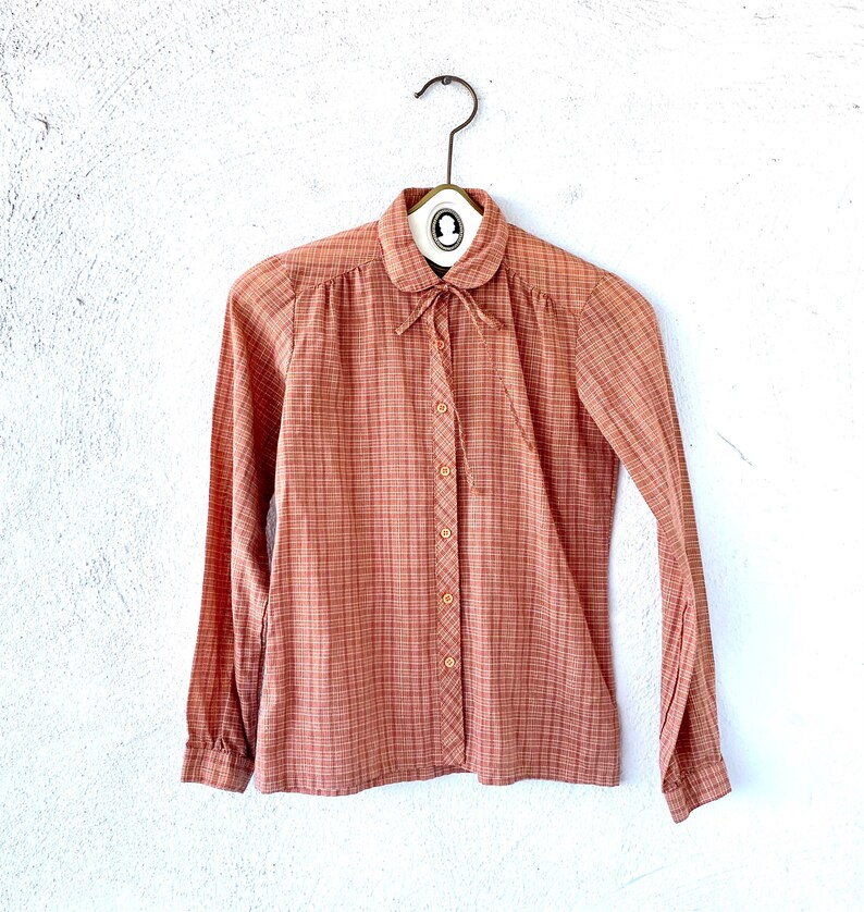 May include: A long-sleeved, button-down shirt with a plaid pattern in shades of pink and orange. The shirt has a Peter Pan collar and a bow tie.