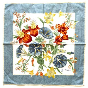 May include: Square silk scarf with a floral design. The scarf features a white center with a colorful arrangement of orange, yellow, and blue flowers. The border is a light blue with a subtle pattern. Signed "Jim Thompson".