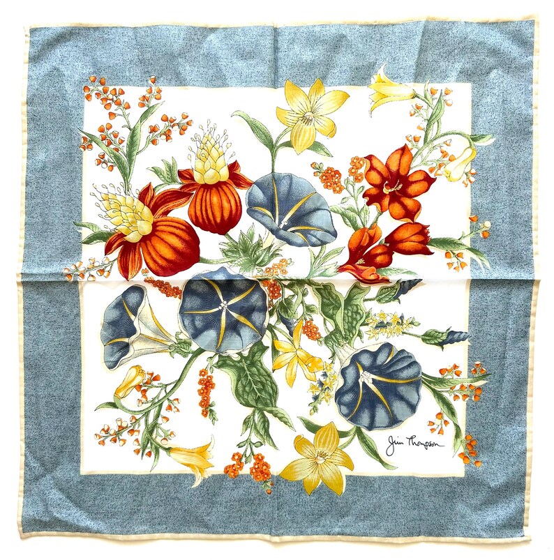 May include: Square silk scarf with a floral design. The scarf features a white center with a colorful arrangement of orange, yellow, and blue flowers. The border is a light blue with a subtle pattern. Signed "Jim Thompson".