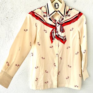 May include: A cream-colored long-sleeved button-up shirt with a red, blue, and white patterned scarf design on the collar. The shirt has a small, repeated pattern of red horseshoes.