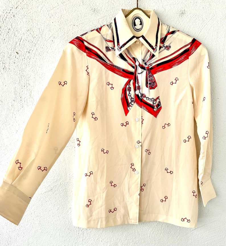 May include: A cream-colored long-sleeved button-up shirt with a red, blue, and white patterned scarf design on the collar. The shirt has a small, repeated pattern of red horseshoes.