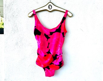 Pode incluir: Um fato de banho de uma peça em rosa vibrante, adornado com um padrão floral arrojado em preto e vermelho. O fato de banho está pendurado num cabide dourado contra uma parede branca. O design apresenta um decote redondo e uma lateral franzida.