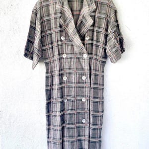 May include: A double-breasted dress featuring a grey, white, and thin red plaid pattern. The garment has short sleeves, a notched lapel collar, and six round white buttons down the front. The fabric has a textured appearance and a straight hem.