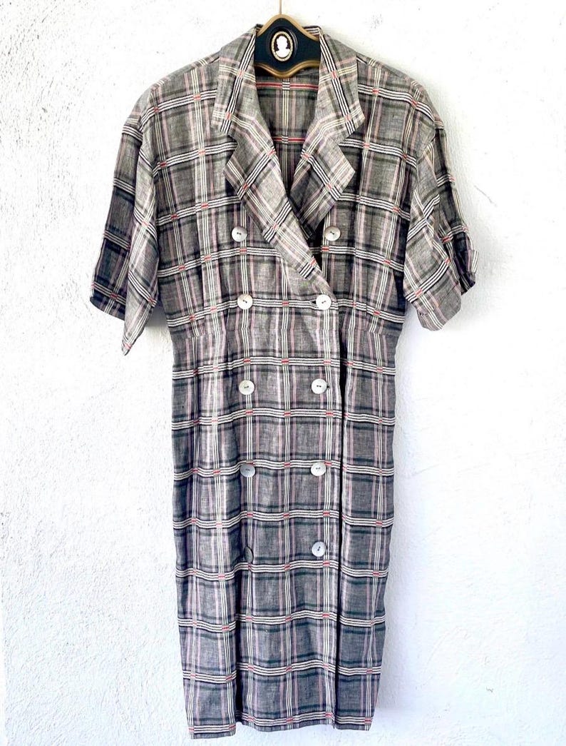 May include: A double-breasted dress featuring a grey, white, and thin red plaid pattern. The garment has short sleeves, a notched lapel collar, and six round white buttons down the front. The fabric has a textured appearance and a straight hem.