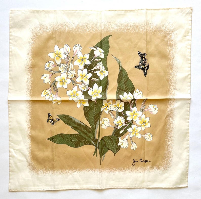 May include: Square silk scarf with a floral design featuring white and yellow flowers, green leaves, and black butterflies. The scarf has a cream-colored border and a tan center. The artist's signature, "Jim Thompson," is visible in the bottom right corner.