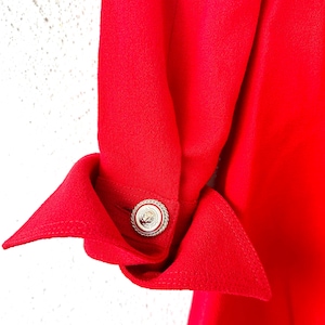 Puede incluir: Primer plano de una manga de prenda roja brillante con un pu&ntilde;o decorativo. El pu&ntilde;o se sujeta con un bot&oacute;n plateado con un dise&ntilde;o detallado. La tela tiene una textura suave y la est&eacute;tica general es elegante.