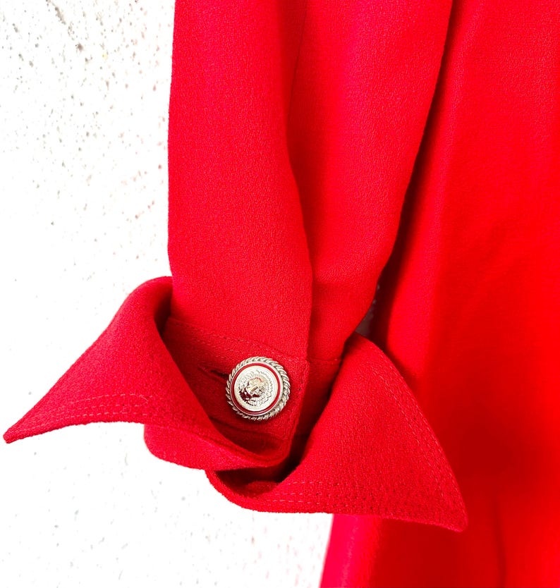 Puede incluir: Primer plano de una manga de prenda roja brillante con un pu&ntilde;o decorativo. El pu&ntilde;o se sujeta con un bot&oacute;n plateado con un dise&ntilde;o detallado. La tela tiene una textura suave y la est&eacute;tica general es elegante.
