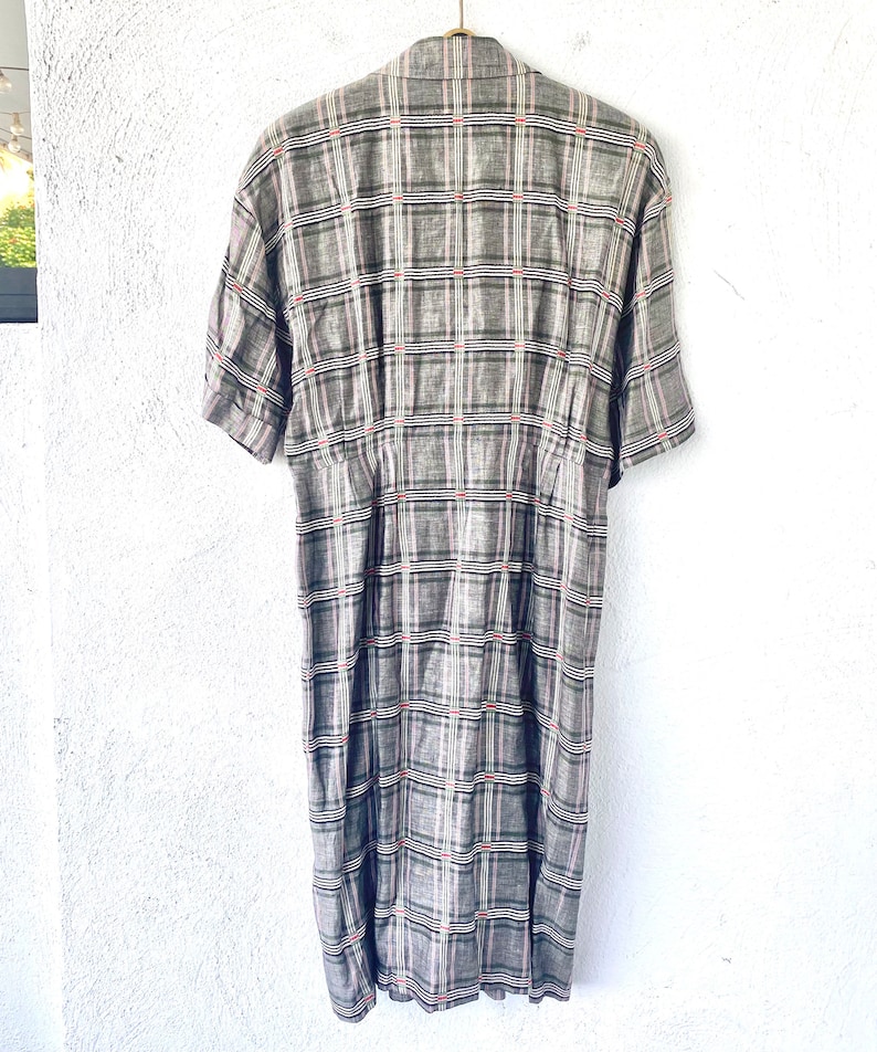 May include: A short-sleeved dress featuring a grey, black, and white plaid pattern with thin red accent lines. The garment has a collared neckline, a button-down front, and a fitted waistline that transitions into a straight skirt silhouette.