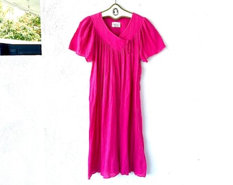Könnte beinhalten: Ein leuchtend pinkfarbenes Vintage-Kleid mit kurzen, flatternden Ärmeln hängt an einem Kleiderbügel. Das Kleid hat einen V-Ausschnitt und einen fließenden, knielangen Rock. Der Stoff wirkt leicht und strukturiert, mit vertikalen Linien. Das Kleid wird vor einer weißen Wand präsentiert.