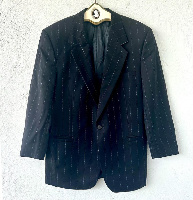 Peut inclure: Un blazer noir avec un motif discret de fines rayures blanches, une fermeture &agrave; un bouton, des revers crant&eacute;s et des manches longues. Le v&ecirc;tement est pr&eacute;sent&eacute; sur un cintre avec un d&eacute;tail cam&eacute;e au col, affichant une coupe structur&eacute;e.