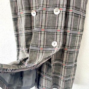 May include: A double-breasted garment featuring a grey, white, and black plaid pattern with thin red accents. The piece is constructed from a textured fabric and includes three large, round, off-white buttons on the front, with a grey lining visible.