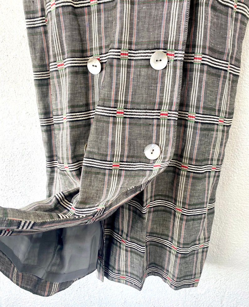 May include: A double-breasted garment featuring a grey, white, and black plaid pattern with thin red accents. The piece is constructed from a textured fabric and includes three large, round, off-white buttons on the front, with a grey lining visible.