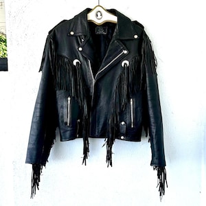May include: Black leather jacket with fringe detailing. The jacket features a classic biker style with a notched collar, a zipper closure, and silver-tone hardware. The fringe is along the sleeves and across the back.