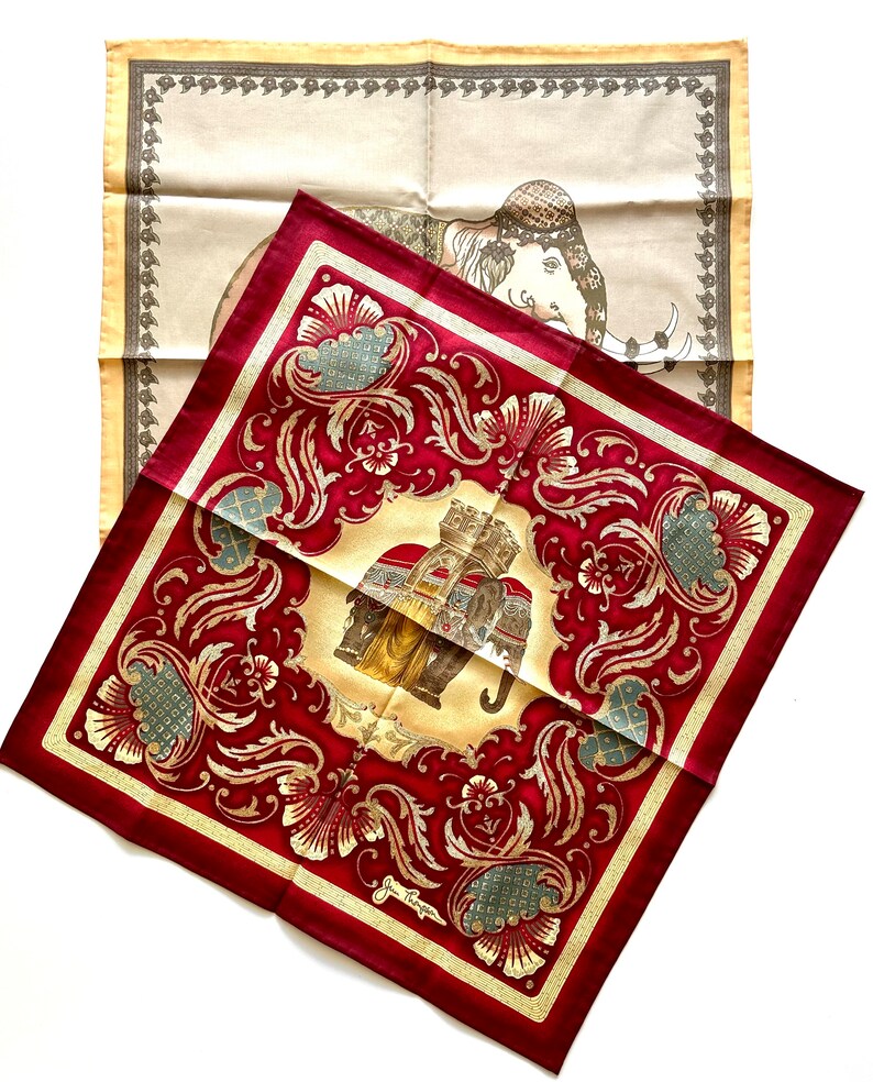 May include: Two silk scarves with elephant designs. One scarf is a deep red with gold floral patterns and an elephant illustration. The other is a cream color with a border and an elephant illustration. Both scarves are square.