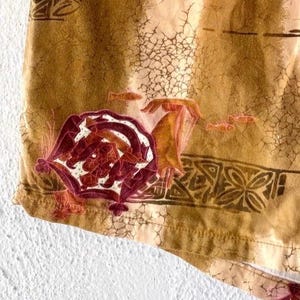 May include: Close-up of a section of fabric with a warm, earthy color palette. The design features a large, ornate floral motif in shades of burgundy and white, with smaller fish and geometric patterns. The fabric has a textured, aged appearance.
