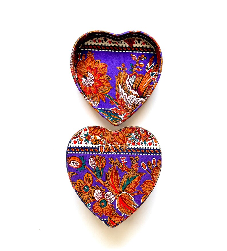 May include: A purple heart-shaped box with a floral pattern in shades of orange, green, and white. The box is open, revealing the inside of the box.