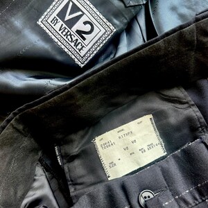 Peut inclure: Un blazer noir &agrave; fines rayures avec une &eacute;tiquette V2 BY VERSACE cousue dans la doublure int&eacute;rieure. Une &eacute;tiquette rectangulaire blanche est fix&eacute;e &agrave; la ceinture int&eacute;rieure, indiquant le mod&egrave;le K17XPX, la taille 52R et divers codes de production.