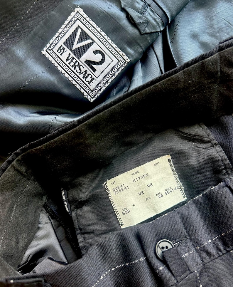 Peut inclure: Un blazer noir &agrave; fines rayures avec une &eacute;tiquette V2 BY VERSACE cousue dans la doublure int&eacute;rieure. Une &eacute;tiquette rectangulaire blanche est fix&eacute;e &agrave; la ceinture int&eacute;rieure, indiquant le mod&egrave;le K17XPX, la taille 52R et divers codes de production.