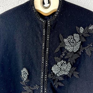 Puede incluir: Una chaqueta de lana negra con ribete de cuentas y dise&ntilde;o floral. Las flores est&aacute;n hechas de cuentas blancas y est&aacute;n bordadas en la chaqueta.